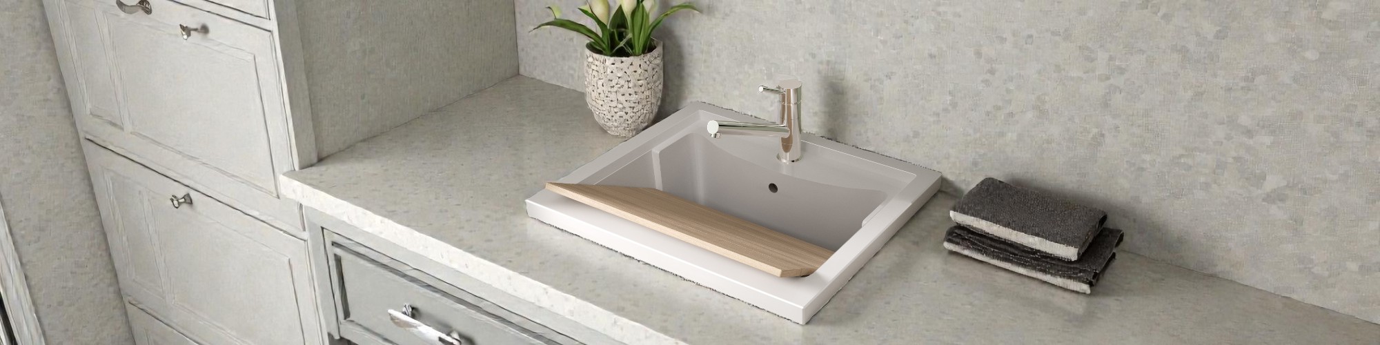 Lavabo moderno empotrado con tabla lavapanni de madera, grifo cromado, planta decorativa y toallas oscuras sobre una encimera de baño clara.